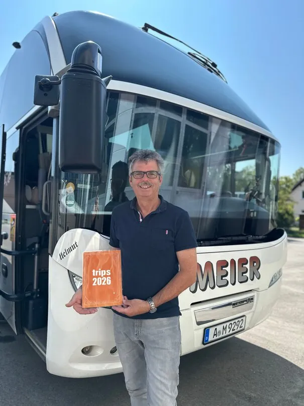 Helmut Haas steht lächelnd vor seinem Reisebus und hält das Reiseprogramm in der Hand.